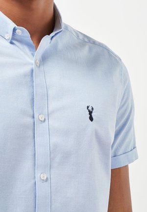 Camisa de manga corta de color azul claro en tela de algodón con cuello abotonado. Presenta un logo de ciervo bordado en negro en el lado izquierdo del pecho.