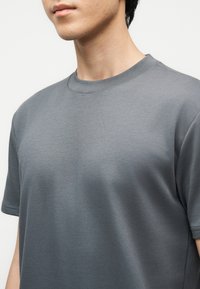 Inimese lähivaade, kes kannab halli lihtsat lühikeste varrukatega ümmarguse kaelusega t-särki heleda tausta ees.