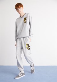Grauer Sweatshirt mit V-Ausschnitt und gelbem aufgesticktem Logo, kombiniert mit passenden grauen Jogginghosen, die dasselbe Logo tragen. Graue Sneaker.