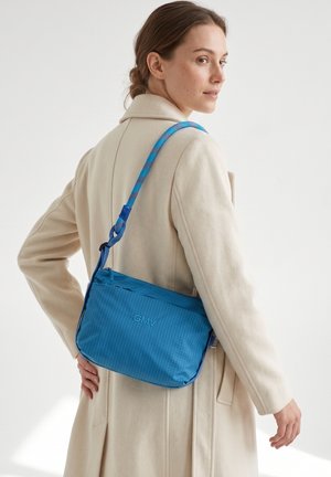 Femme portant un manteau beige avec un sac bandoulière bleu à sangle à motifs et logo GMV, regardant par-dessus son épaule devant un fond uni.