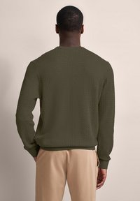 Maglione in verde oliva, lavorato a maglia con texture, collo girocollo e polsini a coste, visto da dietro, abbinato a pantaloni beige chiaro.