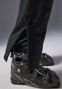 Pantalones de esquí negros con cremalleras laterales, hechos de un tejido duradero, combinados con botas de esquí negras con hebillas ajustables y un acabado texturizado.