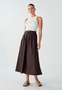 Brauer, gefältete, lange Rock aus einem strukturierten Stoff, kombiniert mit einem taillierten weißen gerippten Tanktop und schwarzen flachen Sandalen.