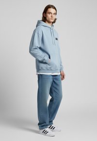 Felpa blu chiaro con tasca anteriore e logo nero, abbinata a jeans blu chiaro e scarpe bianche con strisce nere.