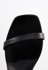 Svart sandal med platt sula, minimalistisk design, slät lädertextur och ett enda tunt band över toppen.