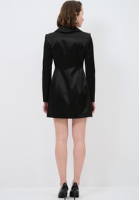 Schwarzes Satin-Kleid mit langen Ärmeln, einschlägigem Kragen und taillierter Passform. Verfügt über einen ausgestellten Rock und eine glatte Textur. Schwarze High Heels runden den Look ab.
