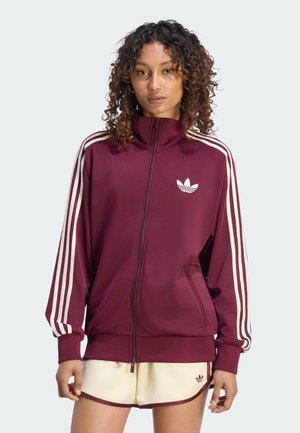 Kobieta ubrana w bordową kurtkę sportową Adidas z białymi paskami oraz kremowe szorty z bordowym wykończeniem, stojąca na jednolitym tle.