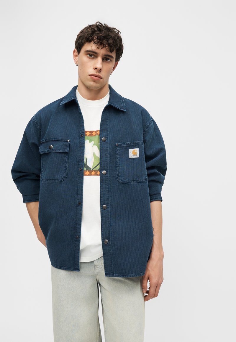 Junger Mann trägt ein marineblaues Cordhemd mit Knöpfen über einem weißen T-Shirt mit floralen Motiven und hellgrauen Jeans, steht vor weißem Hintergrund.