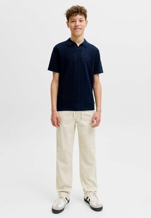 Adolescente con cabello rizado que viste una camiseta polo texturizada azul oscuro, pantalones crema con cordón y zapatillas blancas, de pie frente a un fondo liso.