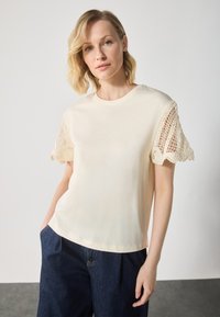 Cream-colored t-shirt with short lace-trimmed sleeves, rounded neckline, and a relaxed fit, paired with dark blue wide-leg jeans.
