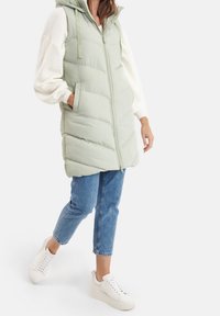 Jasnozielona, puchowa kamizelka z zamkiem z przodu, kapturem i kieszeniami. Połączona z niebieskimi dżinsami i białymi trampkami, z teksturowanym białym swetrem.