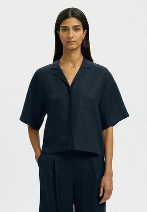 Kvinde med langt, mørkt hår iført en løs, marineblå kortærmet skjorte med knapper og matchende bukser, stående mod en ensfarvet lys baggrund.