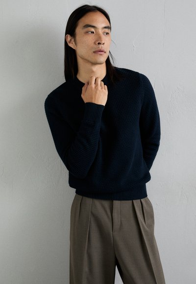 Marineblauer Strickpullover mit strukturierter Oberfläche, Rundhalsausschnitt und gerippten Bündchen, kombiniert mit grauen, gefalteten Hosen. Einfarbig, lässiges Design.