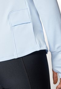 Ljusblå träningsöverdel med sidficka, med en mjuk textur, rundad nederkant och långa ärmar, kombinerad med svarta leggings.