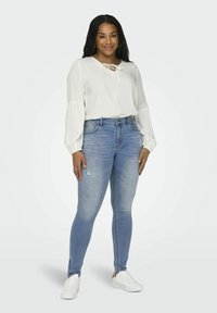 Biela blúzka s dlhým rukávom a šnurovaním, v kombinácii s svetlomodrými priliehavými džínami s poškodenými detailmi a zipsami na členkoch, doplnená bielymi teniskami.