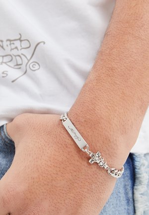 Hõbedane ketiripats, millel on graveeritud ristkülikukujuline plaat ja lillemustrid, tekstureeritud lingid, kantud heleda nahatooni käel.