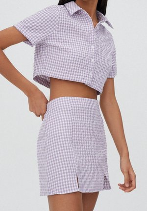 Femme portant une chemise courte boutonnée en vichy lilas et blanc et une mini-jupe taille haute assortie avec de petites fentes sur le devant.
