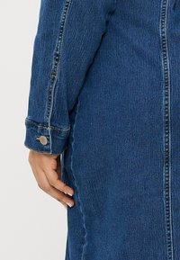 Jaqueta de ganga azul-escura com uma superfície ligeiramente texturizada, apresentando um fecho de correr e um botão de pressão na manga. Detalhe em close da manga e do tecido.