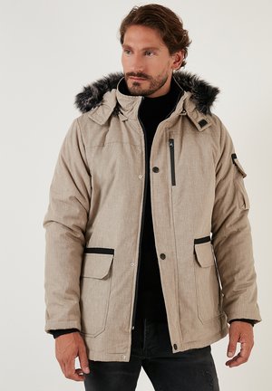 Chaqueta aislante beige con capucha forrada de piel, cierre frontal con cremallera, dos bolsillos delanteros y detalles en negro. Tejido texturizado, corte relajado y capucha desmontable.