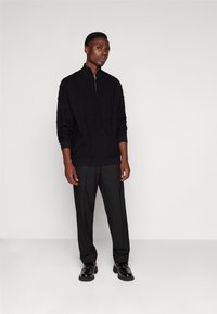 Black knit zip-up sweater with cable pattern, worn with tailored black trousers and black shoes. Model standing against a light backdrop.