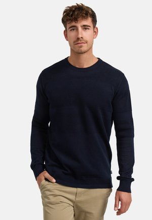 Pullover blu navy con texture, colletto rotondo e maniche lunghe, caratterizzato da un motivo a righe orizzontali e polsini a costine. Indossato con pantaloni kaki.