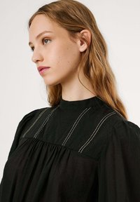 Zwarte blouse met textuur, featuring een hoge hals, pofmouwen en decoratieve zilveren stiksels aan de voorkant. Eenvoudig en elegant ontwerp.