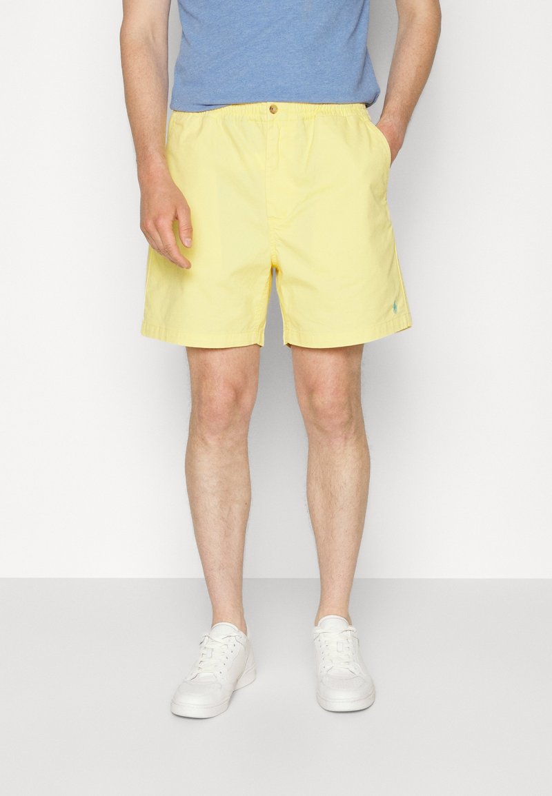 Homme portant un short jaune clair, un t-shirt bleu et des baskets blanches, debout devant un fond uni avec une main dans la poche.