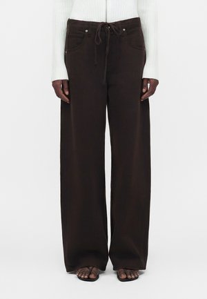 Pantalon droit jambes larges marron foncé avec taille à cordon, porté avec un haut blanc côtelé à manches longues et des sandales noires à bout ouvert.