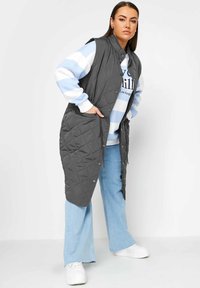 Gilet grigio trapuntato con colletto alto, chiusure a pressione e tasche laterali, indossato sopra una felpa a strisce blu e bianco e jeans di denim.
