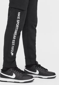 Pantalones deportivos negros con texto blanco que dice "NIKE SPORTSWEAR EST.1972", combinados con zapatillas negras que presentan el logotipo de Nike Swoosh en blanco.