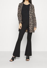 Kobieta ubrana w długi płaszcz w leopardzi wzór, czarną dopasowaną górę, rozkloszowane spodnie i czarne, odkryte czubki wysokie obcasy, stojąca na tle białym.