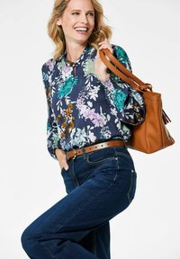 Blumenbluse mit marineblauem Hintergrund, verziert mit grünen, lila und orangen Mustern. Kombiniert mit dunkelblauen Jeans und einer braunen Handtasche.