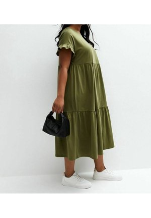 Vrouw in een olijfgroene gelaagde midi-jurk, witte sneakers, die een kleine zwarte handtas vasthoudt tegen een effen witte achtergrond.