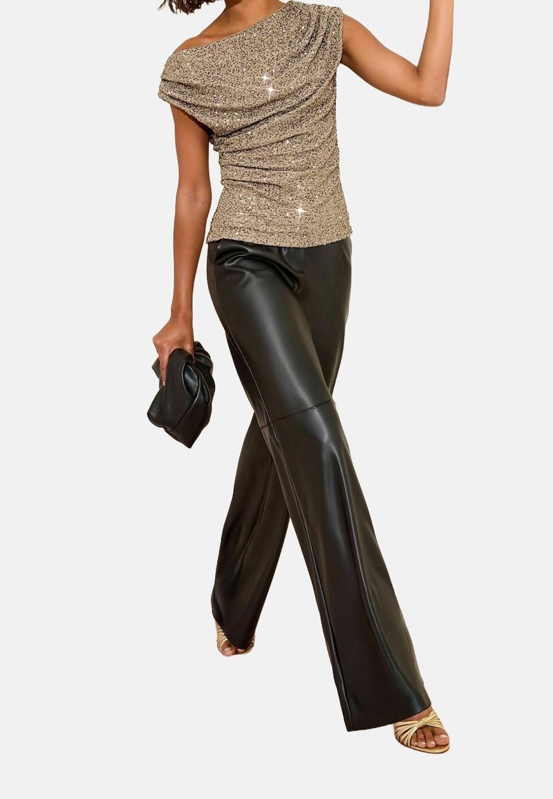 Femme portant un haut beige scintillant asymétrique, un pantalon en cuir noir, tenant une pochette noire et des sandales beiges à lanières.