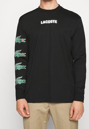 Czarna koszula z długim rękawem z napisem "LACOSTE" na piersi oraz czterema zielonymi logo krokodyla ułożonymi pionowo na lewym rękawie.