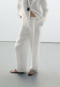 Bílá lněná košile a široké kalhoty, nošeny s černými žabkami s kovovým akcentem. Minimalistický design s uvolněným střihem.