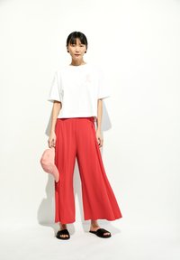Witte cropped t-shirt met een roze letter, rode wijde broek, roze baseballpet en zwarte slide sandalen tegen een witte achtergrond.