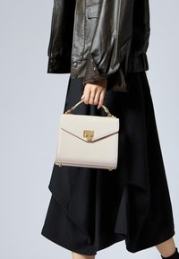 Sac à main crème en cuir à texture crocodile avec des finitions dorées et une anse supérieure, tenu par une main, associé à une jupe noire à couches.