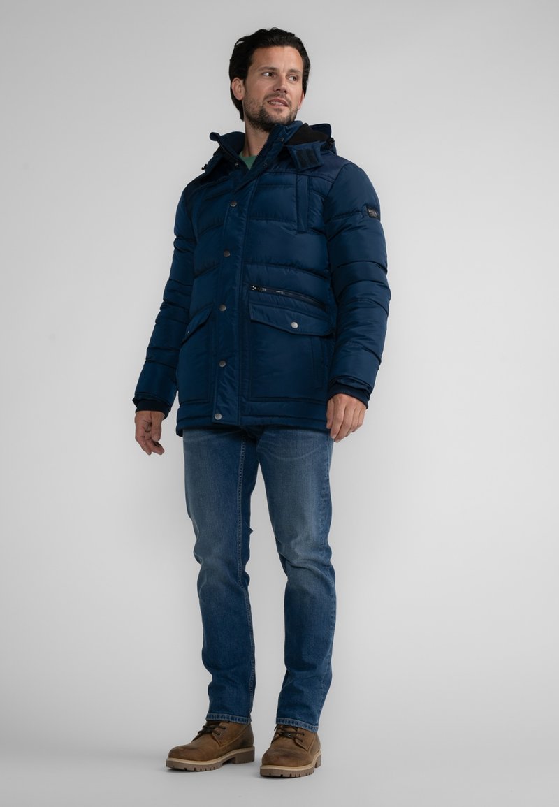 Veste matelassée bleu marine avec capuche, dotée d'une poche zippée, de fermetures à boutons et de poignets élastiques ; portée sur un jean bleu et des bottes crème.