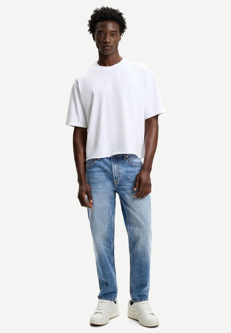 Witte, oversized katoenen t-shirt met korte mouwen, gecombineerd met lichtblauwe, rechte denim jeans. Schoeisel bestaat uit witte sneakers.