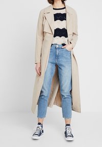 Frau trägt einen beigen Trenchcoat, einen schwarz-weißen gemusterten Pullover, blaue hochtaillierte Jeans und marineblaue High-Top-Sneaker.