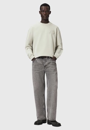 Mand stående med hænderne i lommerne iført en lys cremefarvet sweatshirt med et lille logo og falmede grå straight-leg jeans.