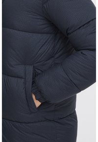 Navy pufferjack met een geruit patroon. Kenmerken zijn onder andere een gestikt textuur, elastische manchetten en zijzakken. Zacht, geïsoleerd materiaal.
