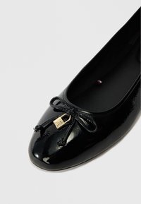 Ballet flat en cuir verni noir avec un bout arrondi, ornée d'un accent nœud et d'un détail en métal doré à l'avant. Texture lisse et brillante.