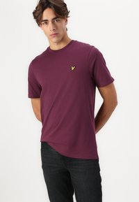T-shirt court maroon en tissu doux, avec une petite broderie d'oiseau vert sur la poitrine et un col rond.
