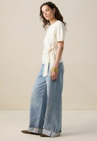 T-shirt crème à manches courtes avec un détail noué à l'avant, associé à un jean en denim bleu clair à jambe large avec un ourlet légèrement effiloché.