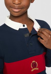 Granatowa i czerwona koszulka polo z białym kołnierzykiem, z guzikami i haftowanym logo. Tekstura wydaje się gładka i nieco prążkowana.