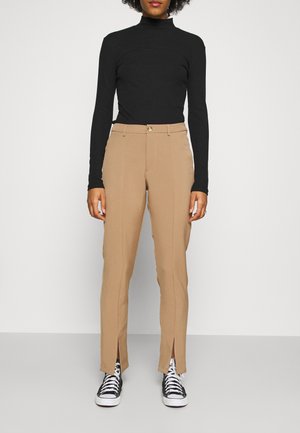 Camisola preta de gola alta com riscas, calças beiges ajustadas com ranhuras laterais e ténis pretos. As calças apresentam um fecho frontal por botão e um design fluido.