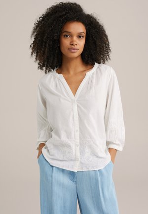 White button-up blouse with a V-neck, 3/4 sleeves, and embroidered detailing at the hem; paired with light blue loose-fitting trousers.