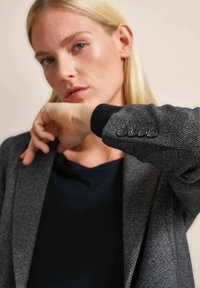 Gray textured blazer featuring black cuffs with button accents, worn over a dark top. The background is a neutral tone, emphasizing the outfit.